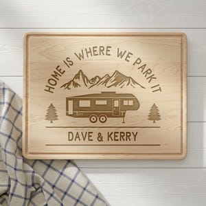 May include: A rectangular wooden cutting board with a carved design. The design includes a camper, mountains, and text that reads "HOME IS WHERE WE PARK IT" and "DAVE & KERRY". Two trees flank the camper.