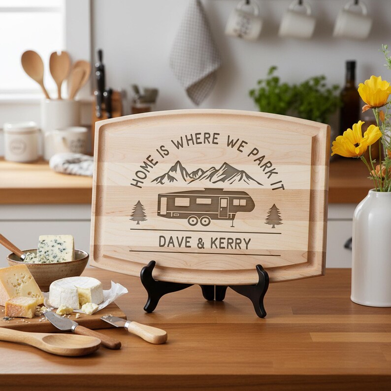 May include: A light-colored wooden cutting board with a decorative design featuring a camper, mountains, and the text "HOME IS WHERE WE PARK IT" and "DAVE & KERRY". The board is displayed on a black stand.