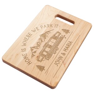 May include: A rectangular wooden cutting board with a handle. The board is engraved with a camper, mountains, and the text "HOME IS WHERE WE PARK IT" and "JOHN & MARIA".