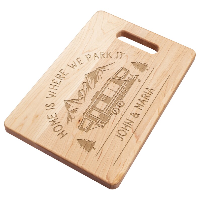 May include: A rectangular wooden cutting board with a handle. The board is engraved with a camper, mountains, and the text "HOME IS WHERE WE PARK IT" and "JOHN & MARIA".