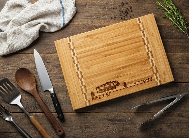 May include: A rectangular bamboo cutting board with a camper design and the words "HAPPY CAMPERS" and "FRED & DEBBIE" engraved on it. Kitchen utensils, including a knife, wooden spoon, and spatula, are arranged around the board.