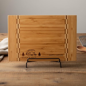 May include: A rectangular bamboo cutting board with a decorative border and a black metal stand. The board features an engraved camper design with the text "GEORGE & HOLLY HAPPY CAMPERS".