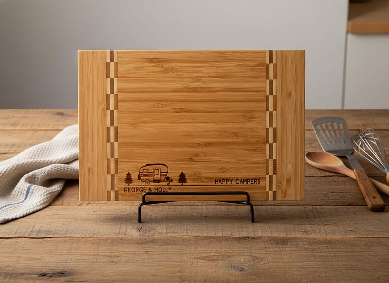 May include: A rectangular bamboo cutting board with a decorative border and a black metal stand. The board features an engraved camper design with the text "GEORGE & HOLLY HAPPY CAMPERS".