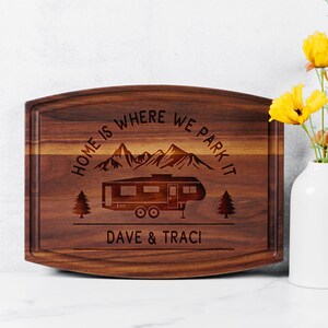 May include: A rectangular wooden cutting board with a dark brown finish. The board features engraved text that reads "HOME IS WHERE WE PARK IT" above a camper illustration and the names "DAVE & TRACI". Two trees and mountain range are also engraved.