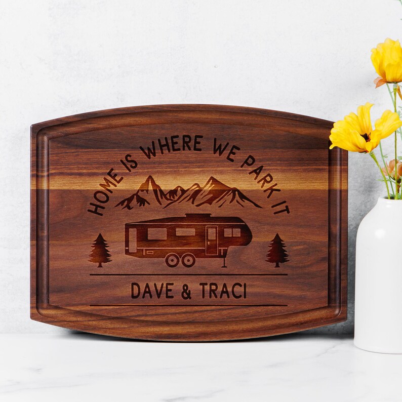 May include: A rectangular wooden cutting board with a dark brown finish. The board features engraved text that reads "HOME IS WHERE WE PARK IT" above a camper illustration and the names "DAVE & TRACI". Two trees and mountain range are also engraved.