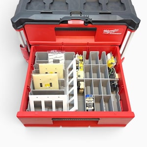 May include: A red and black Milwaukee Packout tool box with a top lid and one open drawer. The drawer contains grey plastic dividers organizing various electrical components, including light switch covers and electrical wall plates.