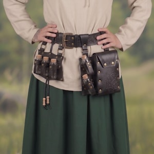 May include: A dark brown leather utility belt featuring an embossed pattern, a brass buckle, and multiple attached components. It includes a rectangular pouch with a star design, brass rivets, and several small glass vials with cork stoppers.