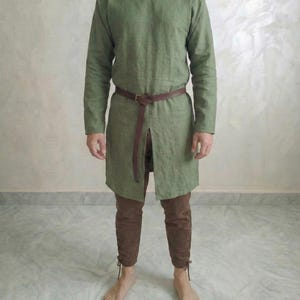 Può includere: Una tunica verde oliva a maniche lunghe con spacco frontale, abbinata a pantaloni marroni e una cintura in pelle marrone. I pantaloni presentano dettagli con lacci alle caviglie. L'outfit evoca un'estetica medievale o storica.