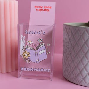 May include: A clear acrylic bookmark holder with a floral design and the text "Sarah's Bookmarks".