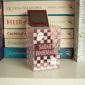 May include: A clear plastic bookmark holder with a pink and white checkered pattern. The holder has the text "Sarah's Bookmarks" in red letters and two white ghost illustrations.