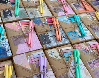 Puede incluir: Una colección de paquetes de papel marrón envueltos con cuerda y decorados con cinta washi colorida y pegatinas. Cada paquete tiene una etiqueta de género diferente, incluyendo "Romance", "Horror", "Ficción" y "Intercambio".