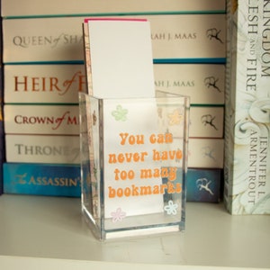 May include: A clear acrylic bookmark holder with a white background and orange text that reads "You can never have too many bookmarks". The holder is decorated with small, colourful flowers.