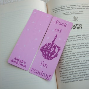 May include: Two pink bookmarks with white polka dots. The bookmark on the left says "Sarah's Book Nook". The bookmark on the right has a purple skeleton hand giving the middle finger and says "I'm reading" and "Fuck off".