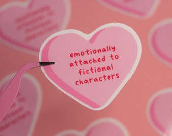 May include: A heart-shaped sticker with a light pink background and a darker pink border. The sticker features the red text "emotionally attached to fictional characters" printed in the center. The sticker is held by a pair of light pink tweezers.