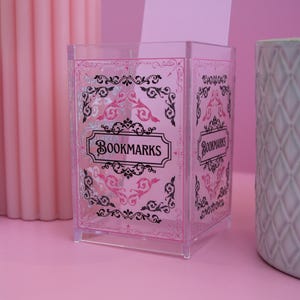 May include: A clear plastic bookmark holder with a pink and black floral design. The holder has the word "BOOKMARKS" printed on the front.
