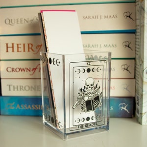 Peut inclure: Un organiseur de bureau en acrylique transparent avec un motif de carte de tarot. La carte est la carte "Le Lecteur", qui est le numéro XI du jeu de tarot. La carte représente un squelette lisant un livre avec une lune croissante et des étoiles en arrière-plan.