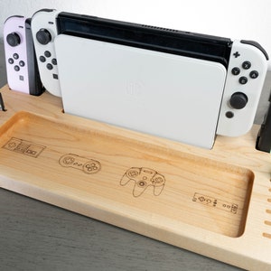 May include: A wooden Nintendo Switch dock with a white Switch console, two controllers, and a game cartridge. The dock has a slot for the console, two slots for controllers, and a slot for a game cartridge. The dock is also engraved with a Nintendo logo and a retro video game controller.