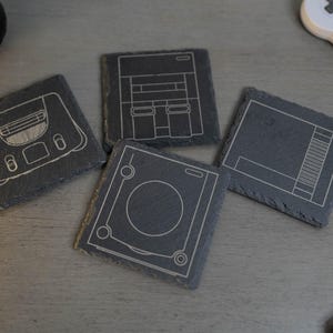 May include: Four square, dark gray slate coasters with white etched designs of classic video game consoles. The coasters include designs of a console, a game system, and a controller. A retro gaming theme.