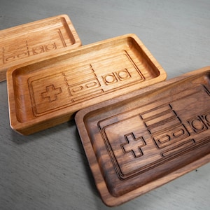 May include: Three wooden trays with a carved design of a classic video game controller. The trays are made of different types of wood, with one being a light brown, one a medium brown, and one a dark brown.