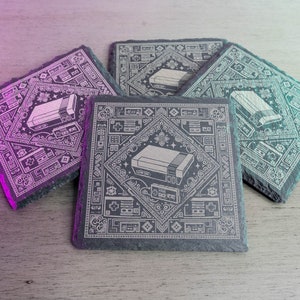 May include: Four black square coasters with a white design featuring a retro video game console and a pixelated pattern. The coasters are arranged in a stack.