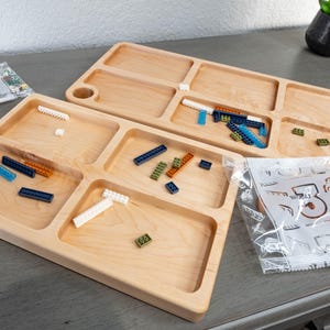 May include: Two wooden trays with compartments for sorting small objects, such as Lego bricks. The trays are made of light wood and have a smooth finish. The trays are filled with various colored Lego bricks, including blue, green, orange, and white.