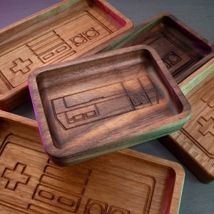 May include: A collection of wooden trays with engraved designs reminiscent of classic video game controllers. The trays are rectangular with raised edges and feature a variety of wood grains and tones, from light oak to dark walnut. The designs include the iconic cross and button layouts.