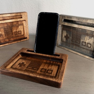May include: Three wooden phone stands with a Nintendo controller design. The stands are made of dark wood and have a slot for a phone. The middle stand has a black phone in it.