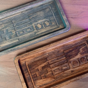 May include: Two wooden trays with a carved design of a classic video game controller. The top tray is a light gray wood with a darker gray stain. The bottom tray is a dark brown wood with a lighter brown stain.