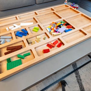 May include: A wooden tray with multiple compartments, filled with colorful Lego blocks. The tray is on a gray table with a black metal base.