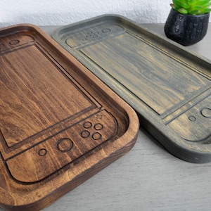 May include: Two wooden trays, one stained dark brown and the other gray, are designed to resemble a game console. The trays have a rectangular shape with rounded corners and feature carved details of buttons and a screen. A small potted plant is in the background.