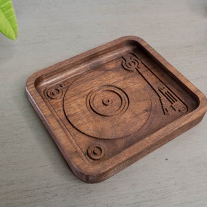 May include: A wooden tray with a carved design of a record player. The tray is dark brown and has a square shape.