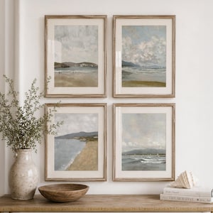 May include: Four framed vertical wall art prints depicting coastal landscapes with sandy beaches, calm ocean waters, and distant hills under cloudy skies. Each print is housed in a light wood frame with a white mat, arranged in a grid pattern.