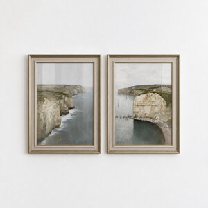 Può includere: Due stampe artistiche da parete incorniciate verticalmente che raffigurano paesaggi costieri con alte scogliere beige, acqua di mare grigio scuro e piccole barche a vela. Ogni stampa è in una cornice metallica chiara con bordo neutro.