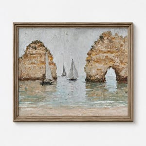 May include: A framed oil painting depicting a coastal scene with two large, textured tan rock formations, one featuring a natural archway. Three white sailboats are positioned on the calm, light blue water, set against a muted gray sky.