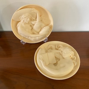 May include: Two white ceramic wall plaques featuring a woman in a reclining pose. One plaque depicts the woman reading a book, while the other shows her wearing a large hat.