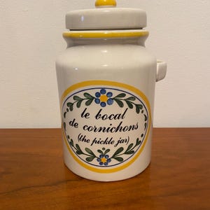 May include: White ceramic pickle jar with a yellow lid and a blue and green floral design. The jar has the text "le bocal de cornichons (the pickle jar)" on the front.