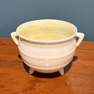 May include: A small, off-white ceramic cauldron-style bowl with a rounded body, a wide opening, and two small side handles. The bowl features three short, stubby legs at the base and has a smooth, glossy finish throughout its exterior surface.
