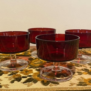 May include: Four vintage ruby red glass dessert bowls with clear stems and bases. The bowls have a wide, shallow design. The glasses are arranged on a patterned tablecloth.