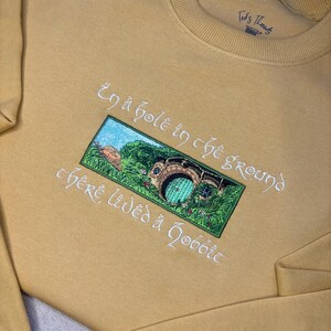 K&ouml;nnte beinhalten: Senffarbenes Sweatshirt mit gesticktem Design. Das Design zeigt ein gr&uuml;nes und blaues Bild eines Hobbit-Lochs mit dem Text "In a hole in the ground there lived a hobbit" in wei&szlig;er Schrift. Der Markenname "Ted's Threads" ist oben rechts aufgedruckt.
