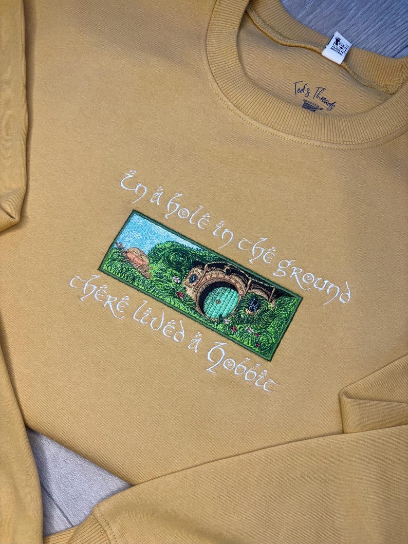 K&ouml;nnte beinhalten: Senffarbenes Sweatshirt mit gesticktem Design. Das Design zeigt ein gr&uuml;nes und blaues Bild eines Hobbit-Lochs mit dem Text "In a hole in the ground there lived a hobbit" in wei&szlig;er Schrift. Der Markenname "Ted's Threads" ist oben rechts aufgedruckt.