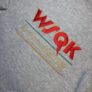Könnte beinhalten: Graues Sweatshirt mit gesticktem rotem Text "WSQK" über "The Squawk 94.5 FM" in Gold. Das Design umfasst blaue und orangefarbene Linien. Ein hölzernes Blumendesign ist in der oberen linken Ecke sichtbar.