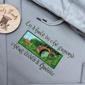 K&ouml;nnte beinhalten: Hellgrauer Kapuzenpullover mit Kordelzug und detaillierter Stickerei. Das Design zeigt ein gr&uuml;nes und braunes Hobbitloch mit dem Text "In a hole in the ground there lived a hobbit" in gr&uuml;ner Schrift.