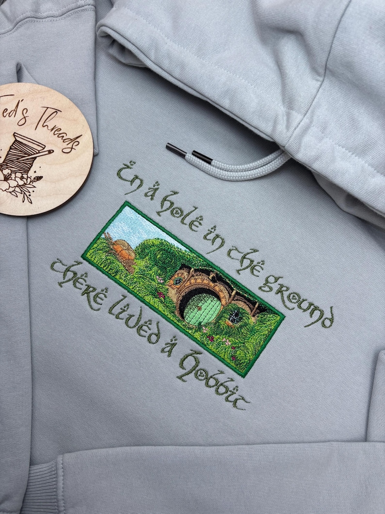 K&ouml;nnte beinhalten: Hellgrauer Kapuzenpullover mit Kordelzug und detaillierter Stickerei. Das Design zeigt ein gr&uuml;nes und braunes Hobbitloch mit dem Text "In a hole in the ground there lived a hobbit" in gr&uuml;ner Schrift.
