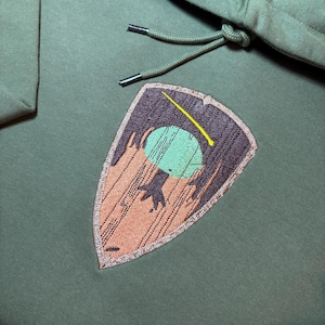 Puede incluir: Sudadera con capucha verde oliva con capucha con cordón. La sudadera tiene un parche en forma de escudo con un diseño bordado de un planeta verde, una estrella fugaz amarilla y elementos marrones y naranjas. El cordón tiene puntas metálicas plateadas.