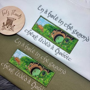 K&ouml;nnte beinhalten: Wei&szlig;e und olivgr&uuml;ne Sweatshirts mit gestickten Kunstwerken. Das Design zeigt eine gr&uuml;ne T&uuml;r in einem grasbewachsenen H&uuml;gel, mit dem Text "In a hole in the ground there lived a Hobbit" in einer stilisierten Schriftart. Ein Holzlogo ist ebenfalls sichtbar.