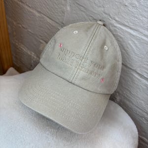 Peut inclure: Casquette de baseball beige avec le texte brodé "support your local library" en fil marron clair. De petits accents de fleurs roses sont brodés près du texte. La casquette est posée sur une surface blanche et moelleuse.