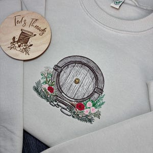 May include: A light gray sweatshirt with an embroidered image of a round wooden door with a small window, surrounded by flowers and greenery. The text "Ted's Threads" is on a wooden tag attached to the sweatshirt.