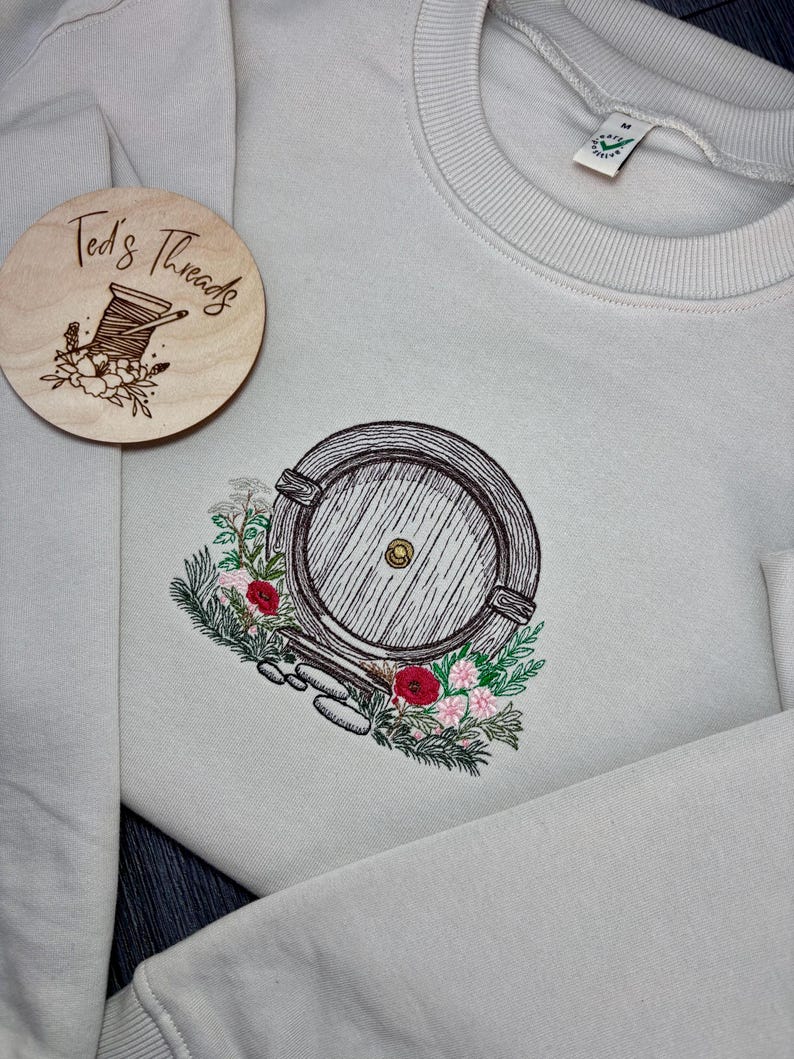 May include: A light gray sweatshirt with an embroidered image of a round wooden door with a small window, surrounded by flowers and greenery. The text "Ted's Threads" is on a wooden tag attached to the sweatshirt.
