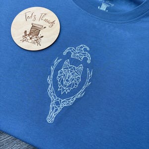 May include: A blue t-shirt with a white geometric wolf design, antlers, and jester hats. A wooden round tag with the text "Ted's Threads" and a spool of thread logo is on the left. The shirt has a crew neck.