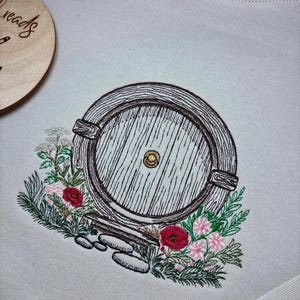 May include: A white sweatshirt with an embroidered image of a round wooden door with a knob, surrounded by green leaves and red and pink flowers.